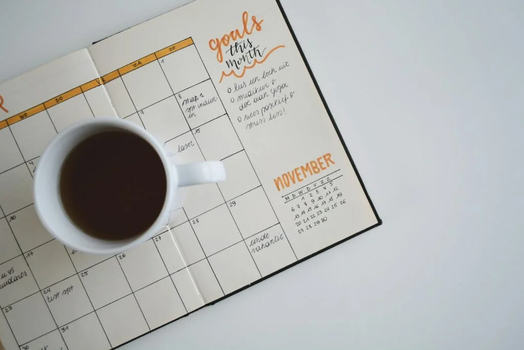 Overcoming Burnout: Rediscovering Joy in Your Work 5 | Mania Africa | maniainc.com A white ceramic mug with coffee on top of a schedule planner - The Role of Organizations in Helping Employees Avoid Burnout - Mania Business