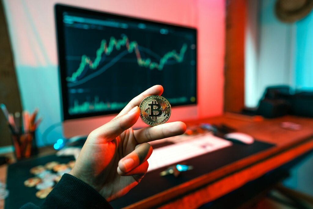 a person holding a Bitcoin coin in front of a computer - A Historical Perspective on Crypto Volatility