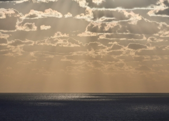 Sunlight streams through clouds over the ocean - A serene, misty horizon where the ocean meets a golden sky, symbolizing the transition between life and the unknown - The Paradox of Life at Death’s Edge: The Gift of Perspective and Lessons from Near-Death Experiences - Mania Lifestyle