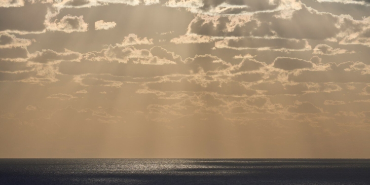 Sunlight streams through clouds over the ocean - A serene, misty horizon where the ocean meets a golden sky, symbolizing the transition between life and the unknown - The Paradox of Life at Death’s Edge: The Gift of Perspective and Lessons from Near-Death Experiences - Mania Lifestyle